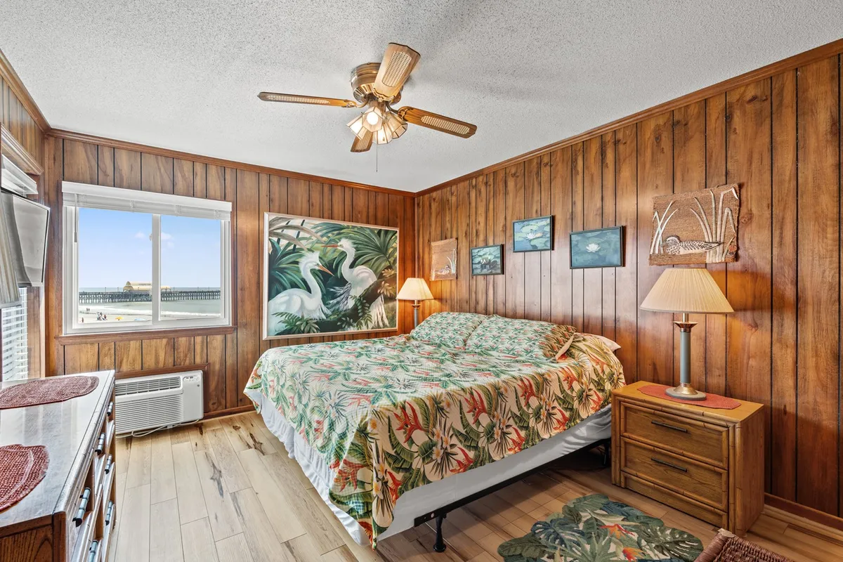 A serene bedroom featuring a large bed with tropical-patterned bedding, wood-paneled walls, artwork on the walls, and a window offering a scenic view. Soft lighting and a plush setup create a tranquil atmosphere ideal for relaxation.