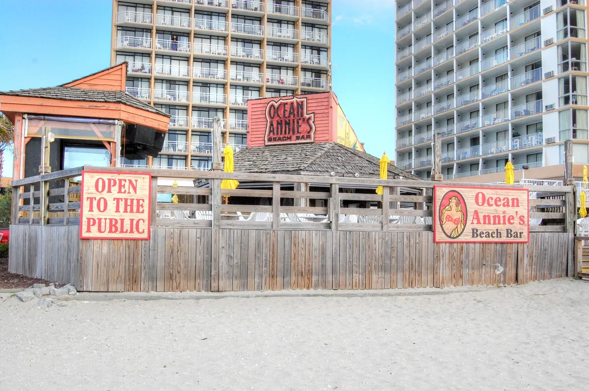 This exterior image shows Ocean Annie's Beach Bar with its prominent sign, wooden fencing, and yellow umbrellas. The setting is cheerful and inviting, with guests socializing and enjoying beachfront dining and live music events just steps from the ocean.