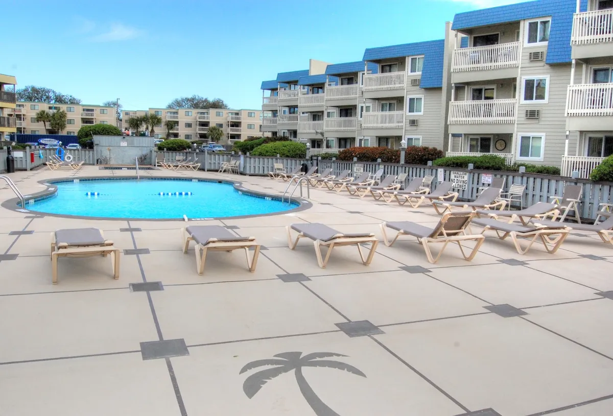 A bright, inviting swimming pool area with several lounge chairs and a relaxed atmosphere, surrounded by the property’s apartments. The setting is ideal for sunbathing, swimming, and mingling with fellow guests.