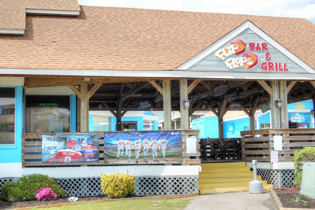 This exterior shot features a lively local bar and grill with a colorful blue exterior and sports-themed banners. People are dining outdoors, chatting with friends, and enjoying the relaxed, festive mood. Large screens display games, and the setting is ideal for group gatherings.