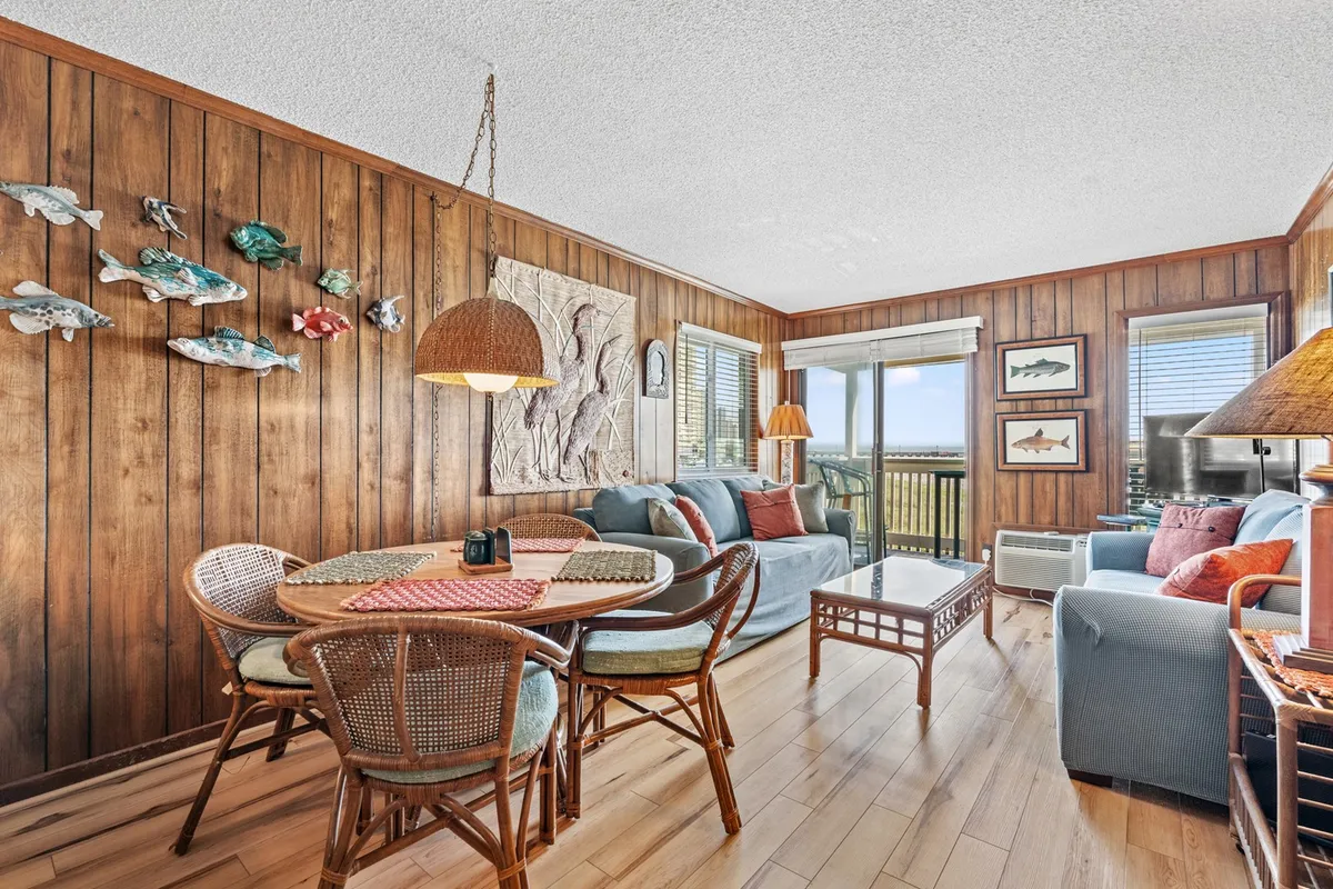 A warm, wood-paneled interior features comfortable couches, decorative fish art, and a dining table with rattan chairs set near a window. The arrangement is cozy and designed for both relaxation and social gatherings, with ample room for guests to interact.