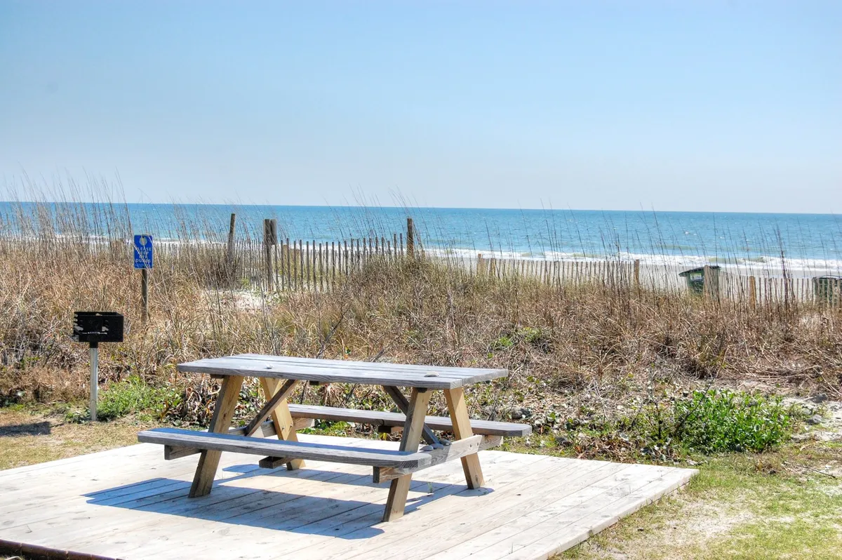 This image features a picnic table set against sweeping ocean views and dune grass, suggesting a peaceful, relaxed seaside atmosphere ideal for quiet reflection or casual gatherings. The scene evokes moments of relaxation and connection with nature.