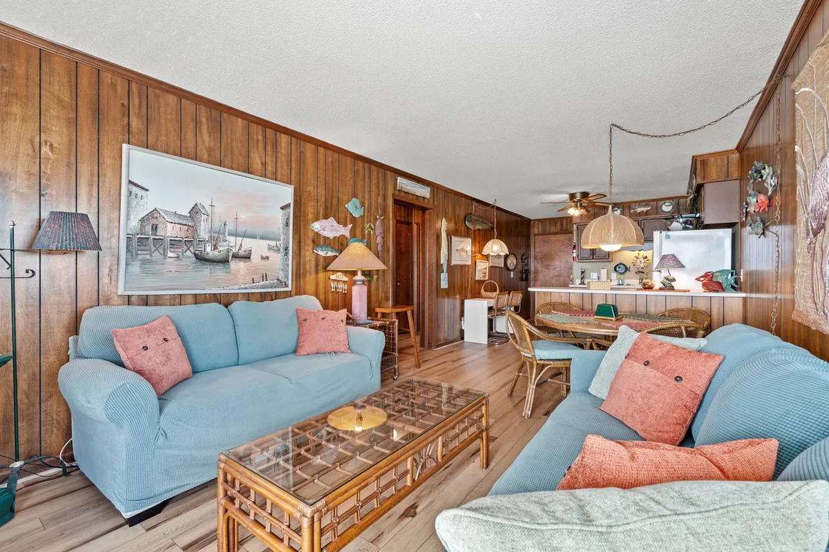 This image shows a casual and welcoming living room featuring a blue sofa, wood paneling, framed artwork, a glass coffee table, and an adjacent dining area. The layout is open, allowing a seamless flow between the living and dining spaces. Natural light streams in, enhancing the inviting atmosphere.