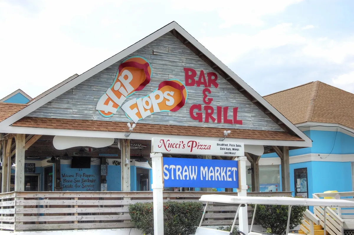 The scene highlights a row of local favorites, including the Flip Flops Bar & Grill, Nucci’s Pizza, and the Straw Market. The area exudes a relaxed and fun vibe with people dining, socializing, and browsing shops, offering a taste of the area's diverse dining and entertainment options.