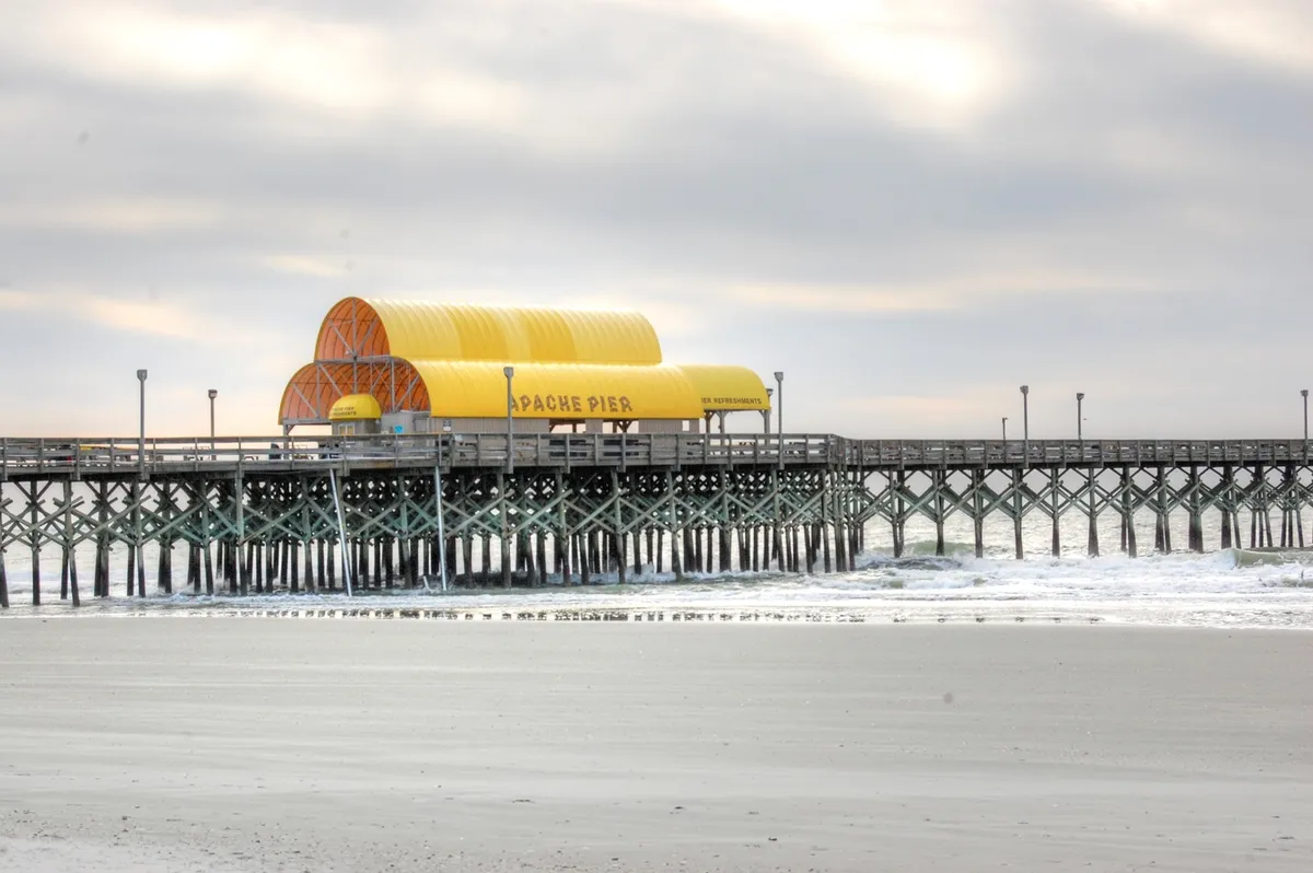 This exterior shot captures the iconic Apache Pier with its distinct yellow roof structure extending over the ocean, gentle waves breaking along the shore. The scene is calm and inviting, illustrating a key local landmark and a place for fishing, strolling, and sunset watching.
