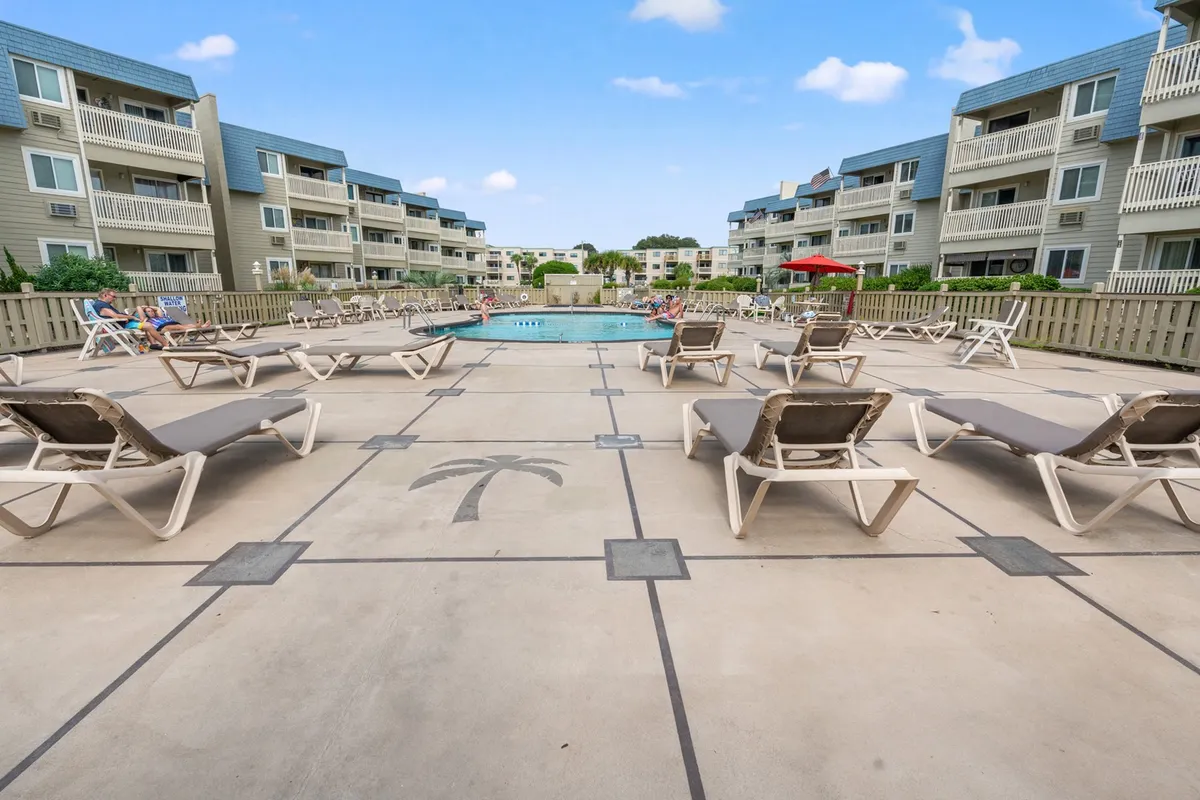 A spacious outdoor swimming pool surrounded by lounge chairs, a pool deck featuring a palm tree design, and several residential buildings in the background. The area is designed for relaxation, socializing, and enjoying the sun.