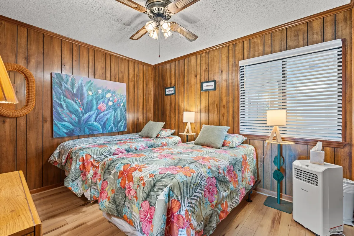 A cheerful bedroom with two twin beds featuring floral bedding, colorful wall art, wood-paneled walls, and a warm-glowing lamp. The room feels bright, welcoming, and perfect for children or friends.