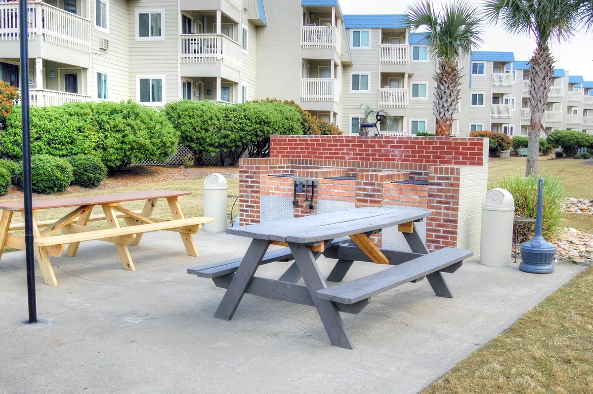 An outdoor grilling and picnic area with a picnic table, a grill, and lush landscaping. The scene invites social gatherings, cookouts, and relaxed meals in a community setting.