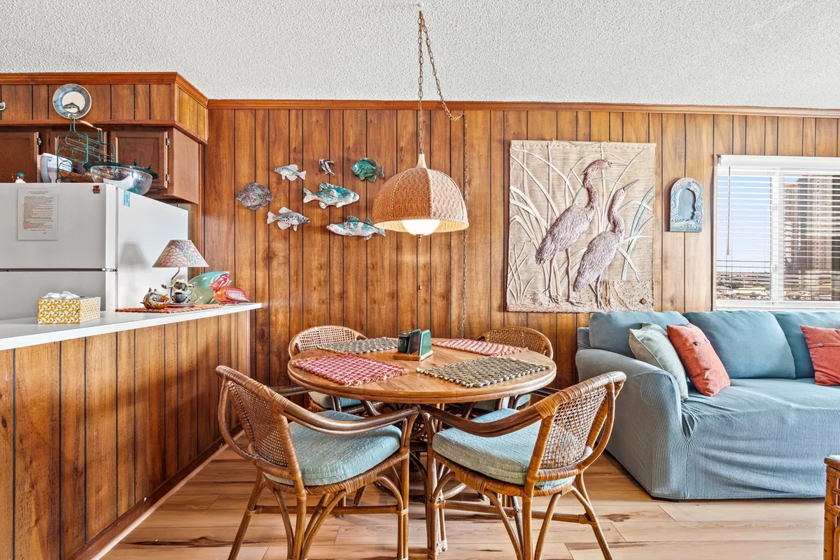 A warm and friendly dining and living area with wood-paneled walls, a round table with woven chairs, a sofa with colorful cushions, and playful fish décor. The space feels intimate and ideal for relaxed evenings or special celebrations.
