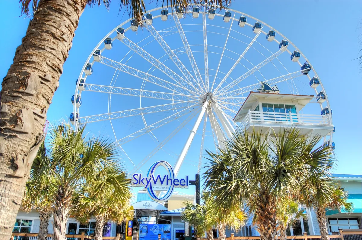This dynamic image showcases the iconic SkyWheel Ferris wheel, framed by palm trees and vibrant signage. The excitement of riding the Ferris wheel and the panoramic views of the coastline are evident, with a lively, family-friendly energy.